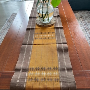 May include: A rectangular table runner with a central mustard yellow panel featuring a geometric pattern. The runner is bordered by brown stripes and white vertical lines. The runner has a fringed edge and is placed on a wooden table.