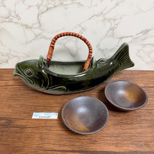Puede incluir: Una fuente de cerámica con forma de pez verde, con un asa tejida y dos cuencos pequeños y redondos. La fuente de pescado tiene un diseño detallado, y los cuencos tienen un esmalte marrón de dos tonos.
