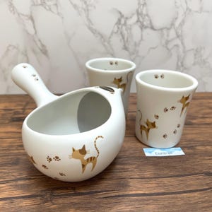 Op de afbeelding: Een witte keramische kat-thema set met een pollepel en twee kleine bekers. De pollepel heeft een handvat en een brede kom. Elk stuk is versierd met bruine kattenillustraties en pootafdrukken.