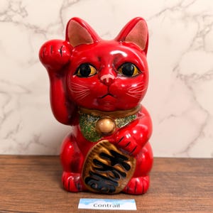 May include: A bright red ceramic lucky cat figurine with a glossy finish. The cat has a raised right paw, large black eyes, and a gold-colored collar with a bell. It holds a gold oval coin featuring black Japanese kanji characters. The word Contrail is printed on a small white label at the base.