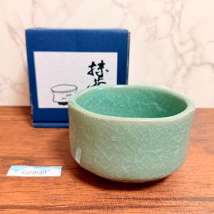 Könnte beinhalten: Eine hellgrüne Keramik-Teeschale mit sichtbarer Craquelé-Glasur. Hinter der Schale steht eine blaue Schachtel mit einem weißen Etikett, das schwarze japanische Kalligrafie und eine Strichzeichnung einer Schale zeigt. Ein kleines Etikett mit Contrail.