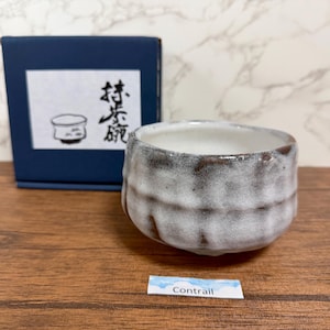 Può includere: Una piccola ciotola in ceramica strutturata con interno bianco ed esterno grigio e marrone. La ciotola è accanto a una scatola blu navy con testo giapponese e un disegno di una ciotola. Una piccola etichetta con la parola "Contrail" è sotto la ciotola.