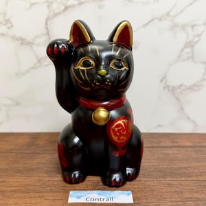 Könnte beinhalten: Schwarze Maneki-neko-Katzenfigur mit erhobener Pfote. Die Katze hat goldene Akzente auf Gesicht und Ohren, ein rotes Halsband mit goldener Glocke und rote und goldene Details am Körper. Auf einer Holzoberfläche.