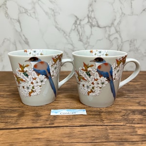 May include: Two ceramic mugs with a bird and floral design. The mugs are light grey with a white handle. Each mug features a blue, brown, and white bird perched on a branch with white flowers. The word "Contrail" is on a small white label.