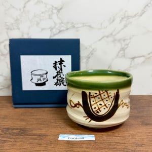 May include: A ceramic matcha bowl with a cream-coloured base and a green rim. The bowl features a brown geometric design and is accompanied by a navy blue box with Japanese text and a matching illustration. The word "Contrail" is on a small label.
