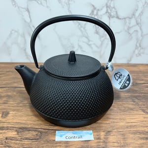 Puede incluir: Una tetera de hierro fundido negro con una superficie texturizada, un asa curva y una tapa con un pequeño pomo. La tetera tiene un pico y está etiquetada con una etiqueta que dice "IWACHU". La palabra "Contrail" está escrita en una pequeña etiqueta.