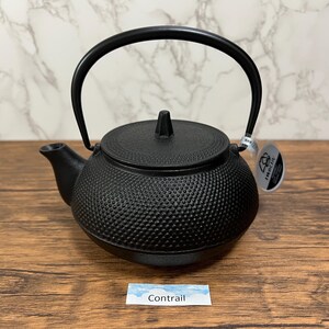 Puede incluir: Una tetera de hierro fundido negro con un asa curva y una superficie texturizada. La tetera tiene un pico y una tapa con un pequeño pomo. La palabra "Contrail" está escrita en una pequeña etiqueta.