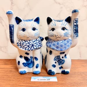 May include: Two white ceramic cat figurines with blue painted details. Each cat has one raised paw, a patterned collar, and blue markings on the body. The left cat features a floral collar, while the right cat has a geometric pattern. Both have Japanese text.