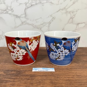 May include: Two ceramic cups featuring bird and floral patterns. One cup is red with white flowers and a bird with blue, black, and brown feathers. The other cup is blue with white flowers and a similar bird. The cups have white interiors.
