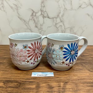 May include: Two ceramic mugs with floral patterns. One mug displays pink and red flowers, the other blue and light blue. Both mugs have a light blue interior and a handle. The word "Contrail" is on a label.