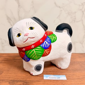 May include: A white ceramic dog figurine with black ears and a curled tail. The dog features a colorful collar decorated with red, green, and blue leaf patterns, a pink nose, and yellow eyes. A small white label with the text Contrail is visible.