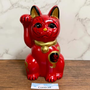 May include: A red ceramic Maneki Neko cat figurine with a raised paw, a gold collar, and gold accents. The cat has black eyes and whiskers. The word "Contrail" is on a small white label at the base.