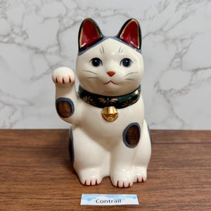 Może przedstawiać: Ceramiczna figurka kota Maneki Neko, biała z czarnymi i szarymi oznaczeniami oraz zielonym kołnierzem ze złotymi akcentami. Kot jest w pozie zapraszającej, z małym złotym dzwoneczkiem. Słowo "Contrail" znajduje się na małej białej etykiecie.