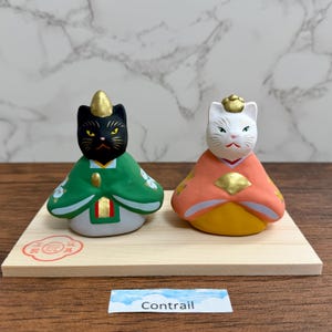 May include: Two ceramic cat figurines, one black and one white, dressed in traditional Japanese attire. The black cat wears a green robe, and the white cat wears an orange robe. Both have gold accents and sit on a wooden base. The word "Contrail" is on a label.