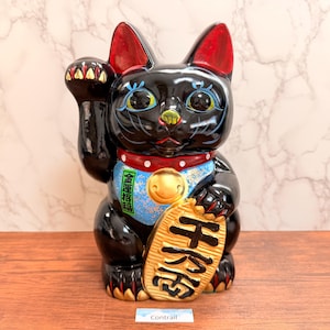 May include: A glossy black beckoning cat figurine with red-lined ears, a red collar with white dots, and a gold bell. The cat holds a gold oval coin with black Japanese characters and features a blue bib with the green text 金運招福.