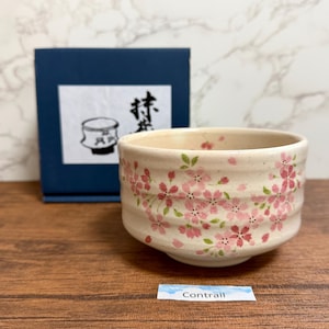 Può includere: Una tazza da tè in ceramica color crema, ornata da motivi di fiori di ciliegio rosa e foglie verdi. La tazza è presentata con una scatola blu con testo giapponese e un disegno. La parola "Contrail" è stampata su una piccola etichetta.