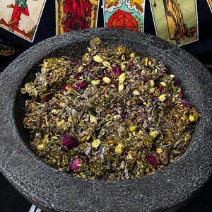 K&ouml;nnte beinhalten: Ein dunkelgrauer Steinm&ouml;rser gef&uuml;llt mit einer Mischung aus getrockneten Kr&auml;utern, Blumen und Knospen. Die Mischung enth&auml;lt violette, rosa, gelbe und gr&uuml;ne Elemente. Tarotkarten sind im Hintergrund zu sehen, was auf ein spirituelles oder rituelles Thema hindeutet.
