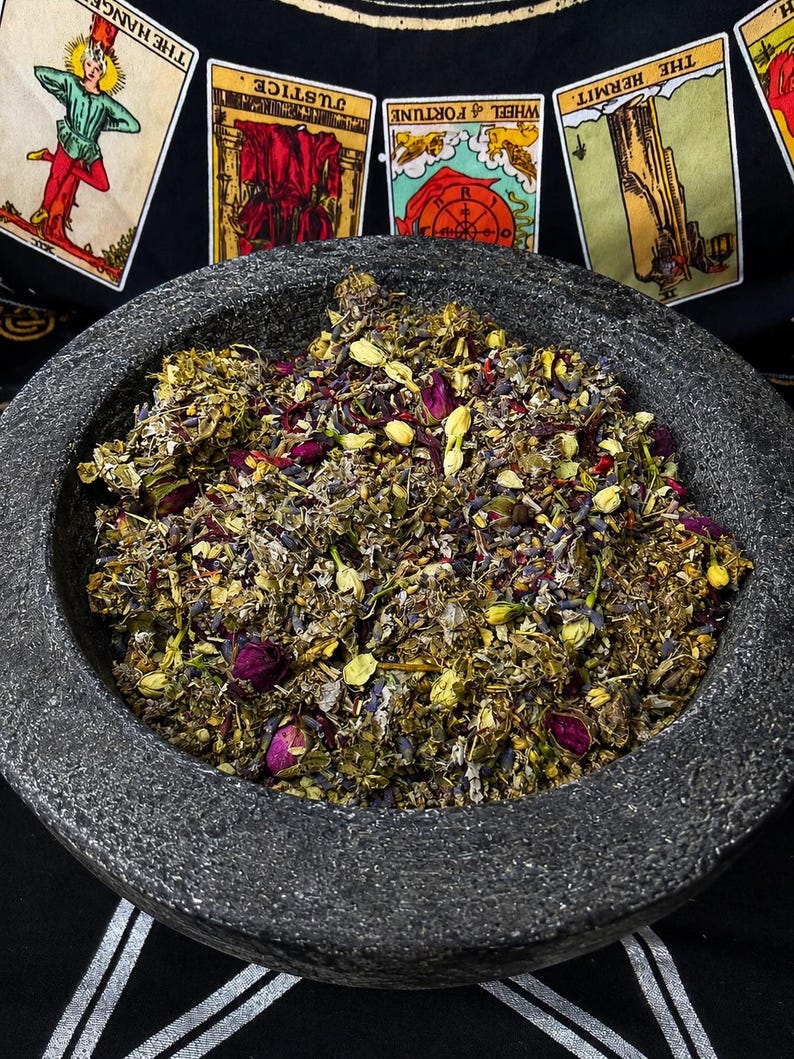 K&ouml;nnte beinhalten: Ein dunkelgrauer Steinm&ouml;rser gef&uuml;llt mit einer Mischung aus getrockneten Kr&auml;utern, Blumen und Knospen. Die Mischung enth&auml;lt violette, rosa, gelbe und gr&uuml;ne Elemente. Tarotkarten sind im Hintergrund zu sehen, was auf ein spirituelles oder rituelles Thema hindeutet.