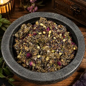 K&ouml;nnte beinhalten: Eine dunkelgraue Steinschale gef&uuml;llt mit einer Mischung aus getrockneten Kr&auml;utern, Bl&uuml;ten und Knospen. Die Mischung enth&auml;lt lila Lavendel, rosa Rosenknospen und gr&uuml;ne Bl&auml;tter. Die Schale steht auf einer Holzoberfl&auml;che, mit einer brennenden Kerze und dekorativen Gegenst&auml;nden im Hintergrund.