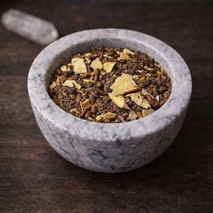 May include: A marble mortar filled with a mixture of herbs and spices. The contents are a mix of brown, tan, and yellow pieces. A clear crystal is in the background. The mortar is on a dark wooden surface.