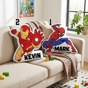 May include: Two superhero-themed throw pillows on a beige sofa. One pillow features Spider-Man in red and blue with the name "MARK". The other pillow depicts Iron Man in red and gold with the name "KEVIN". A colorful blanket and wooden blocks are also present.