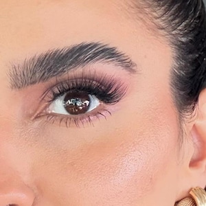 May include: Close-up of a person's face, highlighting the eye with long, dark lashes and pink eyeshadow. The eyebrow is thick and well-defined. A gold earring is visible. The skin has a smooth, even tone.