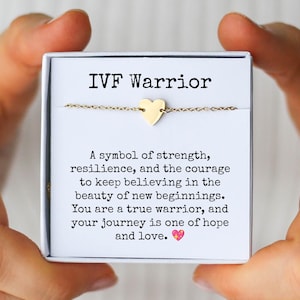 May include: A gold-colored heart charm bracelet presented in a white gift box. The box reads "IVF Warrior" above a message about strength and hope. The bracelet is held by hands.