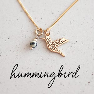 Peut inclure: Un collier doré orné d'un pendentif en forme de colibri avec des détails ajourés. Il comprend également une petite perle ronde à motifs noirs et blancs. Le mot "hummingbird" est écrit en cursive en dessous du collier.