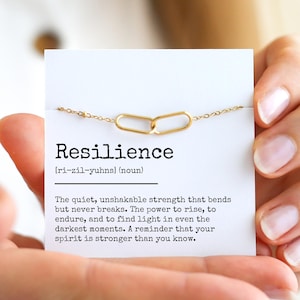 May include: A gold-colored necklace with two interlocking oval links, displayed on a card with the word "Resilience" and its definition. The card is held in a person's hands.