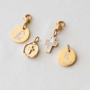 Może przedstawiać: Cztery złote zawieszki z zapięciami na karabińczyk. Jedna zawieszka to okrągły dysk z grawerunkiem ptaka. Druga to zawieszka w kształcie krzyża z inkrustacją z masy perłowej. Trzecia zawieszka to okrągły dysk z literą "K" i datą "11.06.23". Czwarta zawieszka to okrągły wisiorek z krzyżem.