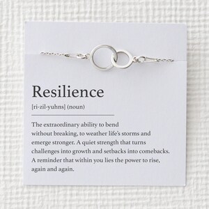 Puede incluir: Un brazalete plateado con dos c&iacute;rculos entrelazados, exhibido en una tarjeta blanca. La tarjeta presenta la palabra "Resiliencia" y su definici&oacute;n, enfatizando la fuerza interior. Una delicada pieza de joyer&iacute;a.