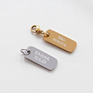 Puede incluir: Dos colgantes rectangulares con texto grabado. Uno es dorado con "R&G FOREVER", el otro plateado con "EMMA 15.5.23". Ambos tienen pequeños bucles para sujetarlos, sugiriendo que son colgantes de joyería o etiquetas.