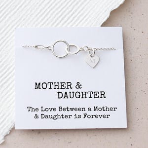 Op de afbeelding: Een zilveren armband met twee in elkaar grijpende cirkels en een hartvormige bedel met de letter "M" op een witte kaart. De kaart leest "MOTHER & DAUGHTER" en "The Love Between a Mother & Daughter is Forever."