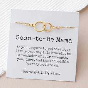 May include: A gold-colored bracelet with two interlocking circles, displayed on a white card. The card reads "Soon-to-Be Mama" with an inspirational message about strength and love. The bracelet is on a light-colored surface.