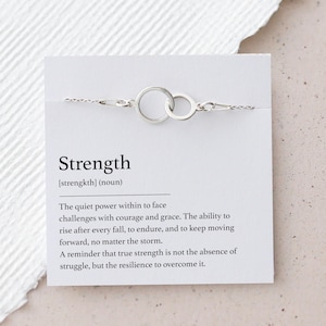 Puede incluir: Una pulsera plateada con dos círculos entrelazados, exhibida en una tarjeta blanca. La tarjeta presenta la palabra "Strength" y su definición. El fondo es una superficie texturizada blanquecina.