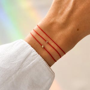 Può includere: Tre braccialetti di filo rosso con piccole perline dorate sono indossati su un polso. I braccialetti sono sottili e delicati, e le perline dorate aggiungono un tocco di brillantezza. Il polso è contro una camicia bianca.