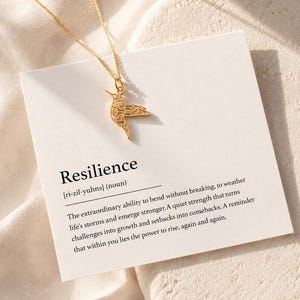 Könnte beinhalten: Eine goldfarbene Halskette mit einem Kolibri-Anhänger. Die Halskette wird auf einer weißen Karte mit der Definition von "Resilienz" präsentiert. Die Karte liegt auf einer strukturierten, cremefarbenen Oberfläche.