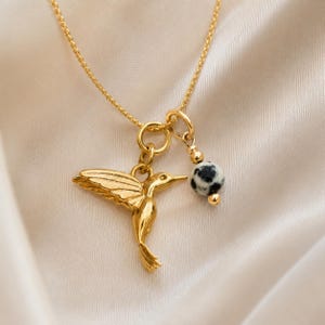 K&ouml;nnte beinhalten: Goldfarbene Halskette mit einem Anh&auml;nger in Form eines Kolibris mit ausgebreiteten Fl&uuml;geln und einem kleinen, runden Anh&auml;nger mit schwarz-wei&szlig;en Punkten. Die Kette h&auml;ngt an einer zarten Kette.