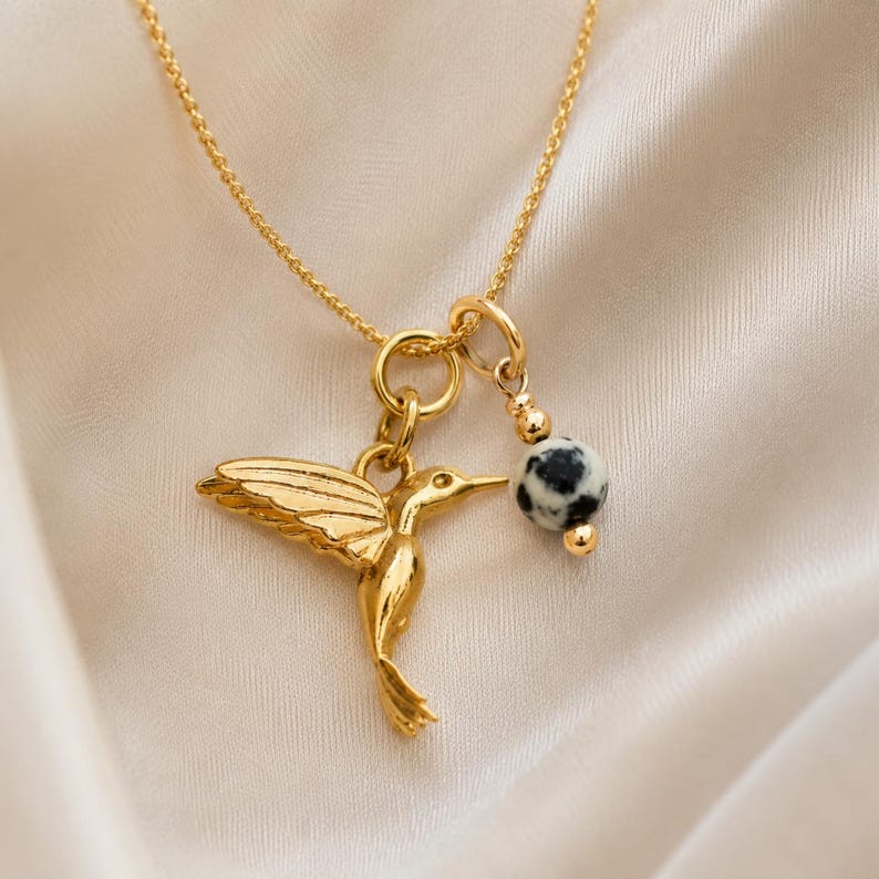 K&ouml;nnte beinhalten: Goldfarbene Halskette mit einem Anh&auml;nger in Form eines Kolibris mit ausgebreiteten Fl&uuml;geln und einem kleinen, runden Anh&auml;nger mit schwarz-wei&szlig;en Punkten. Die Kette h&auml;ngt an einer zarten Kette.