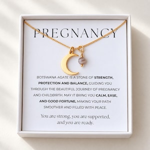 Pode incluir: Um colar de tom dourado com um pingente de meia-lua e uma pequena conta redonda de ágata do Botswana com faixas cinzentas. O colar é apresentado numa caixa branca com o texto PREGNANCY e uma mensagem sobre as propriedades da pedra.