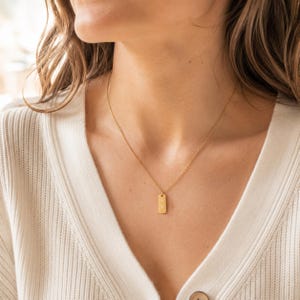 Puede incluir: Un collar de tono dorado con una cadena delicada y un peque&ntilde;o colgante rectangular. El colgante tiene una superficie lisa y pulida con dos diminutas hendiduras o grabados circulares alineados verticalmente en su cara frontal.