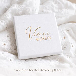 Puede incluir: Una caja de regalo cuadrada de color blanco con una superficie texturizada que presenta el texto Vinci WOMAN impreso en l&aacute;mina dorada en la tapa. La caja est&aacute; colocada sobre un fondo de tela blanca con detalles de bordado ingl&eacute;s.