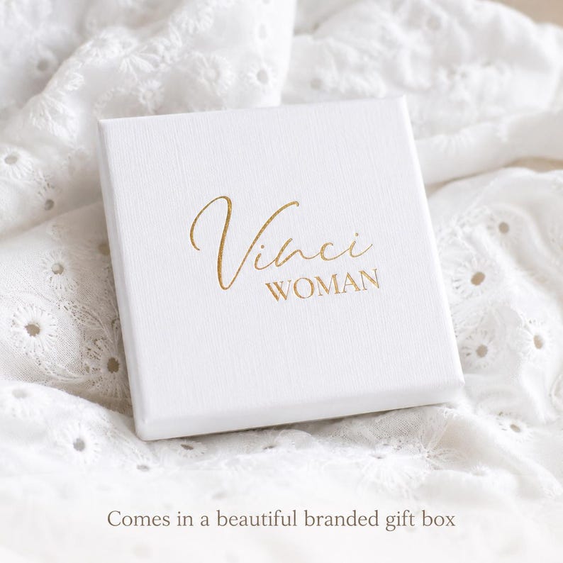 Puede incluir: Una caja de regalo cuadrada de color blanco con una superficie texturizada que presenta el texto Vinci WOMAN impreso en l&aacute;mina dorada en la tapa. La caja est&aacute; colocada sobre un fondo de tela blanca con detalles de bordado ingl&eacute;s.