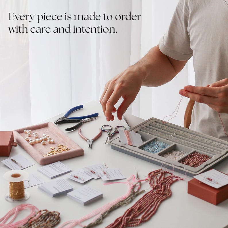 May include: A workspace with jewelry-making supplies. Tools include pliers, scissors, and a tray of beads. The text "Every piece is made to order with care and intention" is visible. Various beads and materials are arranged on a white surface.