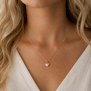 Può includere: Una delicata collana d'oro con un piccolo ciondolo a forma di cuore. Il ciondolo a forma di cuore ha un'iniziale incisa in modo sottile. La collana è indossata su un top bianco, evidenziando il design del gioiello.