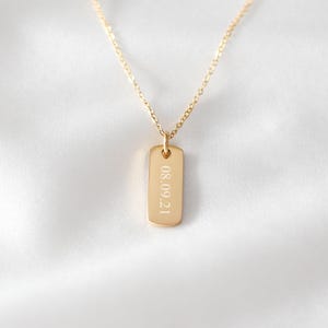 Puede incluir: Un collar de tono dorado con una cadena fina y un peque&ntilde;o colgante rectangular. El colgante est&aacute; grabado con la fecha 08.09.21 en una fuente sans-serif clara. La pieza tiene un acabado pulido y una orientaci&oacute;n vertical sencilla.