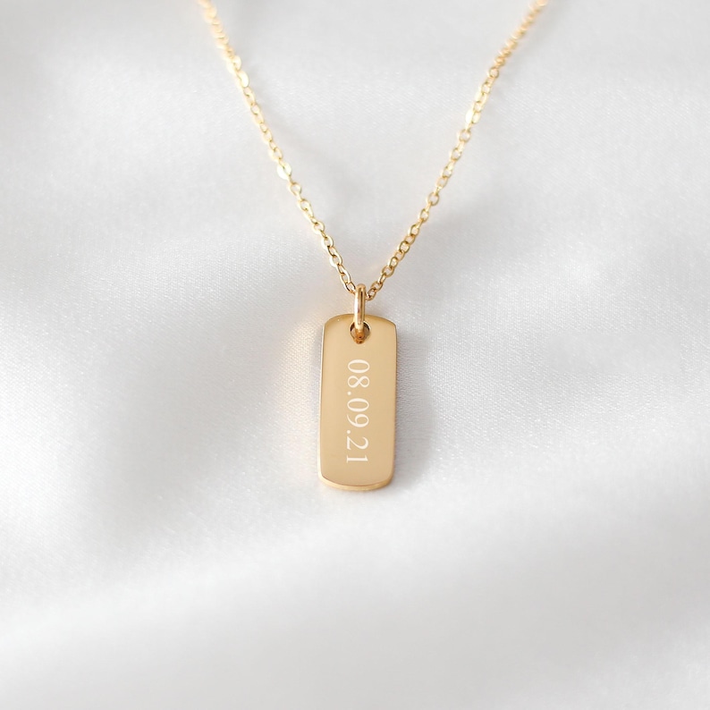 Puede incluir: Un collar de tono dorado con una cadena fina y un peque&ntilde;o colgante rectangular. El colgante est&aacute; grabado con la fecha 08.09.21 en una fuente sans-serif clara. La pieza tiene un acabado pulido y una orientaci&oacute;n vertical sencilla.