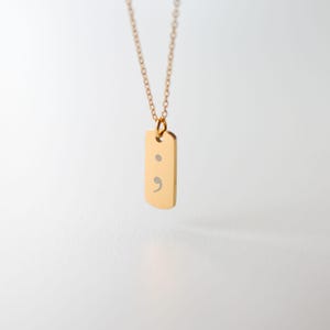 Puede incluir: Un collar de tono dorado con un peque&ntilde;o colgante rectangular de esquinas redondeadas. El colgante tiene grabado un s&iacute;mbolo de punto y coma de color plateado. La pieza cuelga de una delicada cadena de eslabones a juego en tono dorado.