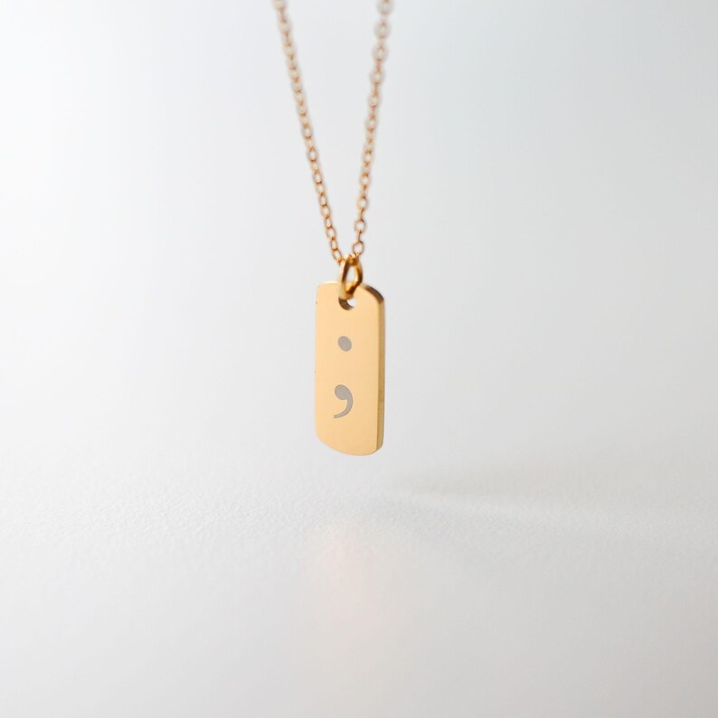 Puede incluir: Un collar de tono dorado con un peque&ntilde;o colgante rectangular de esquinas redondeadas. El colgante tiene grabado un s&iacute;mbolo de punto y coma de color plateado. La pieza cuelga de una delicada cadena de eslabones a juego en tono dorado.
