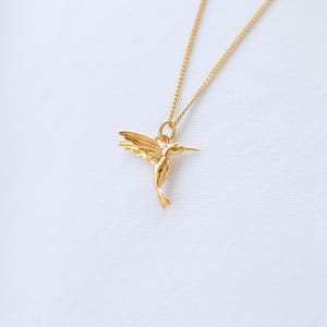 K&ouml;nnte beinhalten: Eine goldfarbene Halskette mit einem Anh&auml;nger in Form eines Kolibris. Der Kolibri befindet sich im Flug, mit ausgebreiteten Fl&uuml;geln. Die Halskette ist zierlich und ebenfalls goldfarben. Der Hintergrund ist ein weicher, wei&szlig;er Stoff.